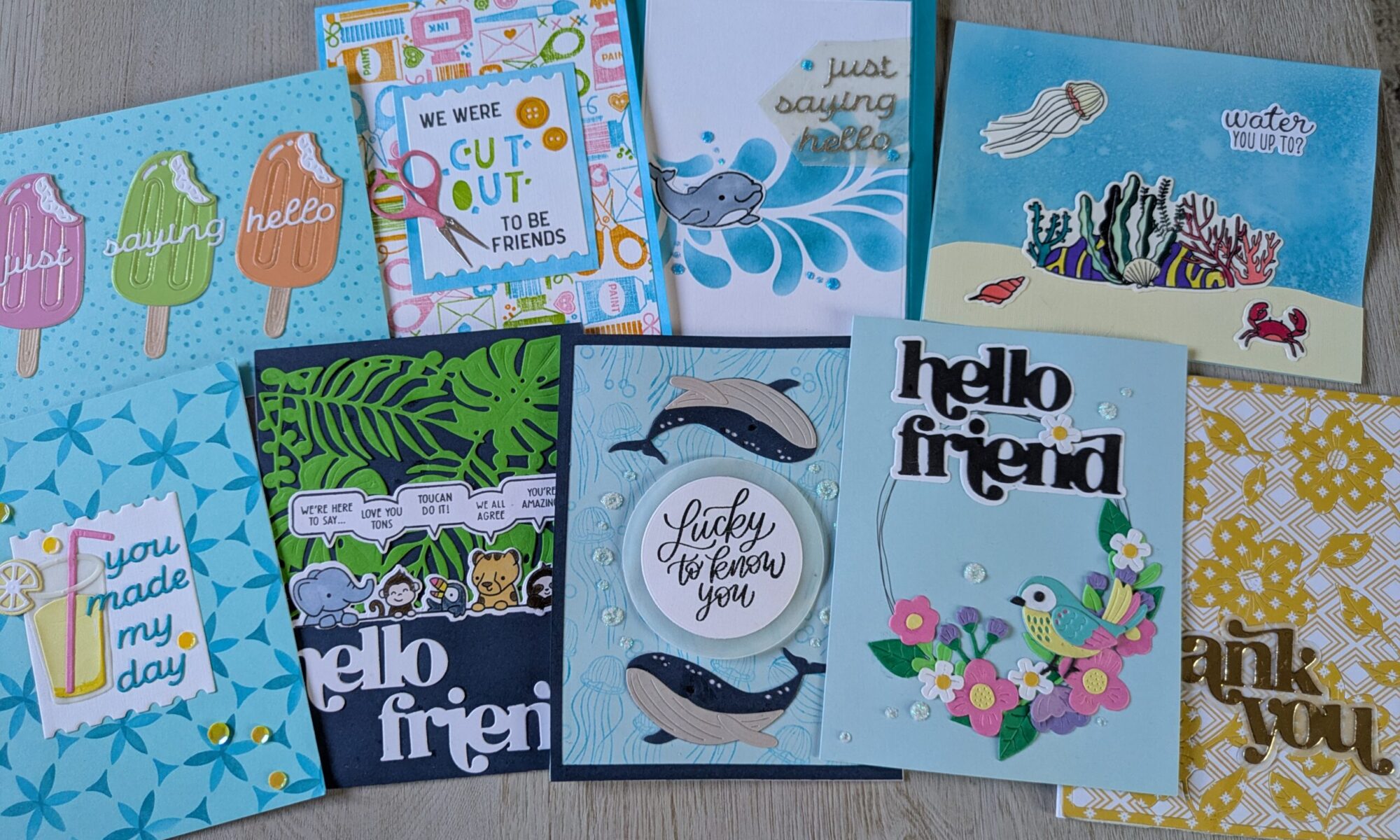 8 overlapping handmade cards are arranged on a wooden table. They are mostly blue and feature cute stamps and die cuts