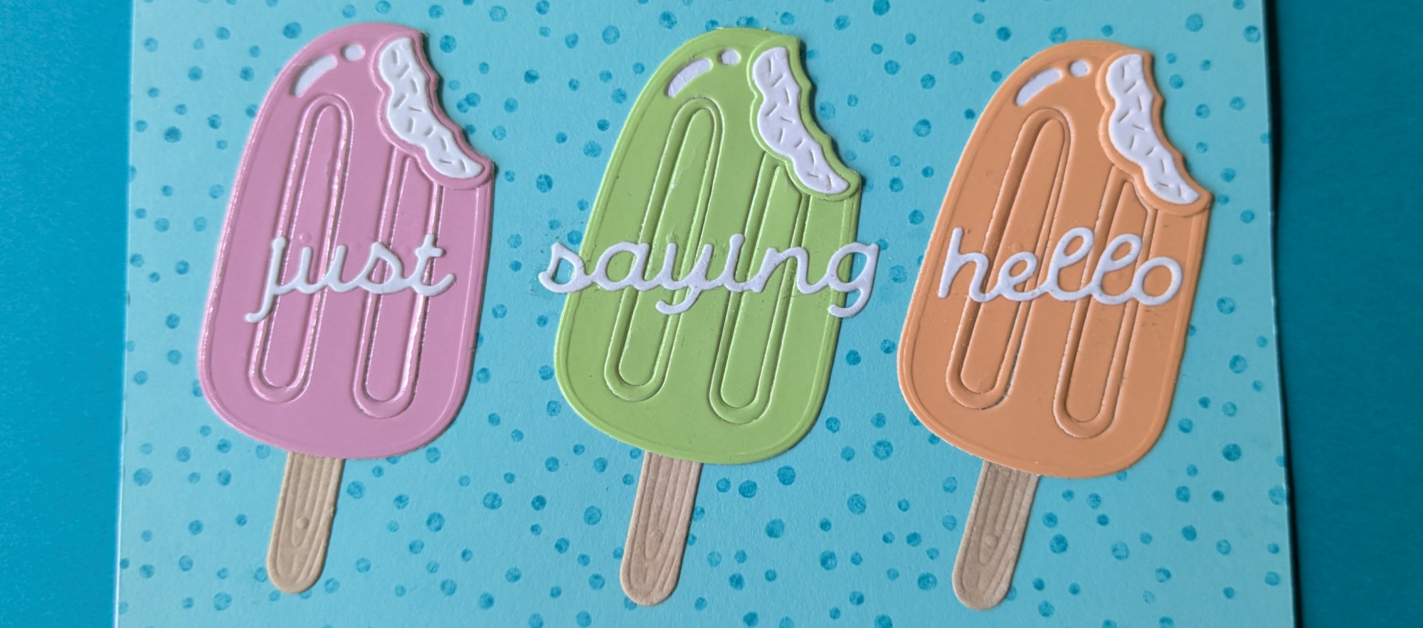 A horizontal card on a dark turquoise background. The base of the card is aqua with speckled turquoise dots. Three creamy popsicles are arranged in a row, one pink, one light green, and one orange. The sentiment "just saying hello" is placed across the popsicles.