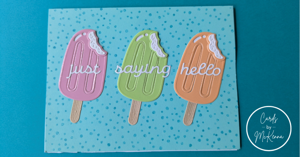 A horizontal card on a dark turquoise background. The base of the card is aqua with speckled turquoise dots. Three creamy popsicles are arranged in a row, one pink, one light green, and one orange. The sentiment "just saying hello" is placed across the popsicles.