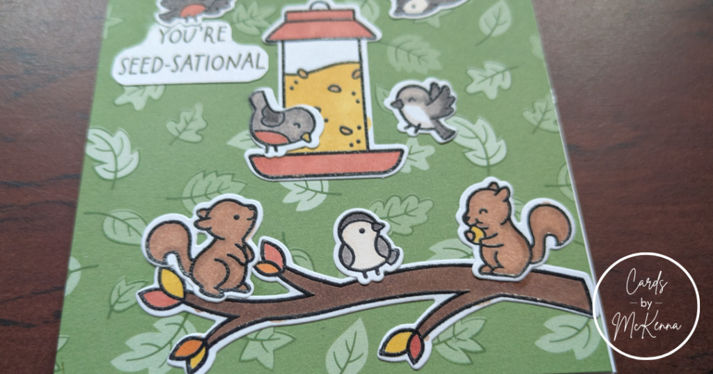 A closer view of the lower half of the same card, focusing on the bottom branch, bottom of the bird feeder, and the two squirrels, one of whom has stolen a seed