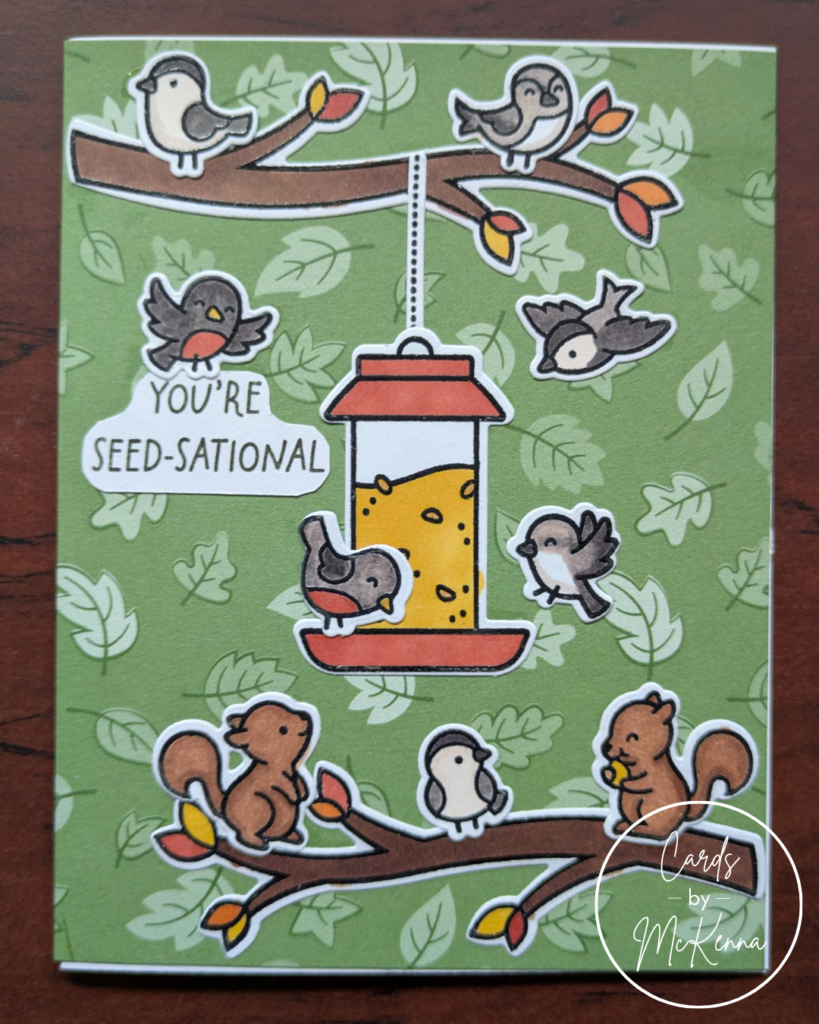 A card with a background of green paper printed with falling leaves. Cutesy birds colored as robins, chickadees, and mockingbirds fly around a hanging bird feeder full of seeds. Some perch on branches with red, orange, and yellow leaves, along with two brown squirrels. One robin holds the sentiment "You're Seed-sational"