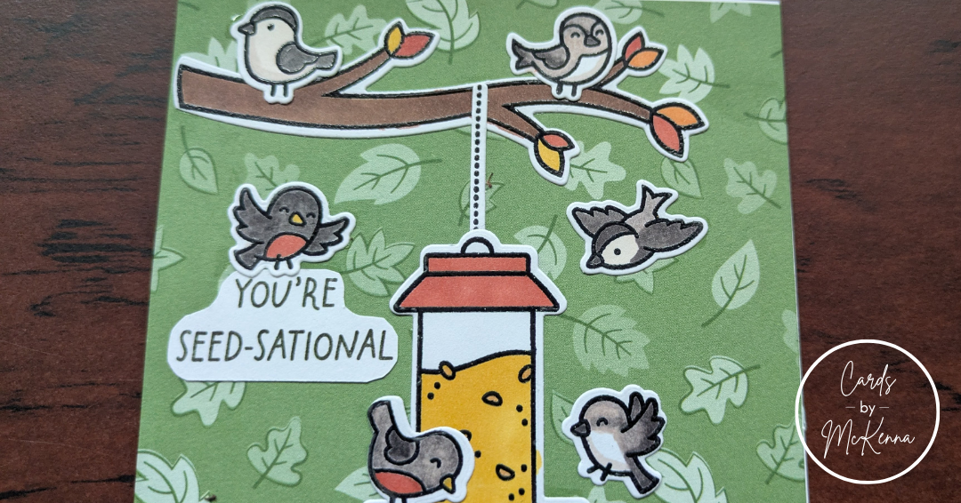 A card with a background of green paper printed with falling leaves. Cutesy birds colored as robins, chickadees, and mockingbirds fly around a hanging bird feeder full of seeds. Some perch on branches with red, orange, and yellow leaves, along with two brown squirrels. One robin holds the sentiment "You're Seed-Sational"