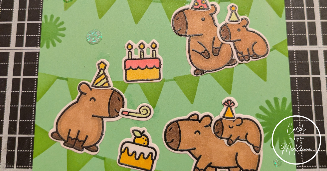A closer view of the capybara birthday card, cropped to focus on the capybaras and their cakes. Scattered between the pennants are dots of holographic glitter glue.
