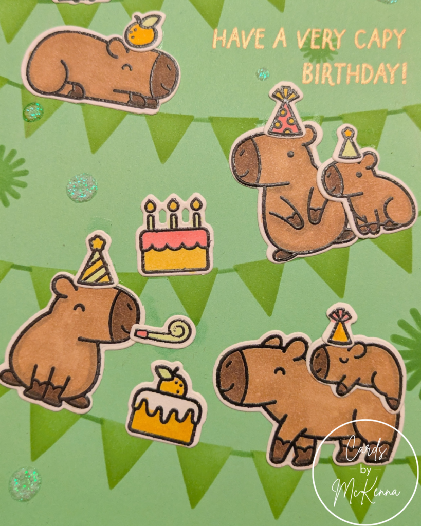 A closer view of the capybara birthday card, cropped to focus on the capybaras and their cakes. Scattered between the pennants are dots of holographic glitter glue.