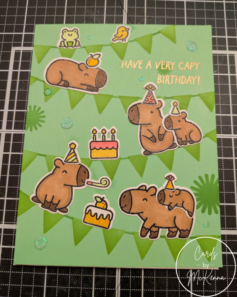 A birthday card with a green paper background with stenciled pennants. Capybaras drawn in a cutesy style perch on the pennant strings with party hats and birthday cake. The sentiment reads "Have a very capy birthday!" in rose gold lettering.